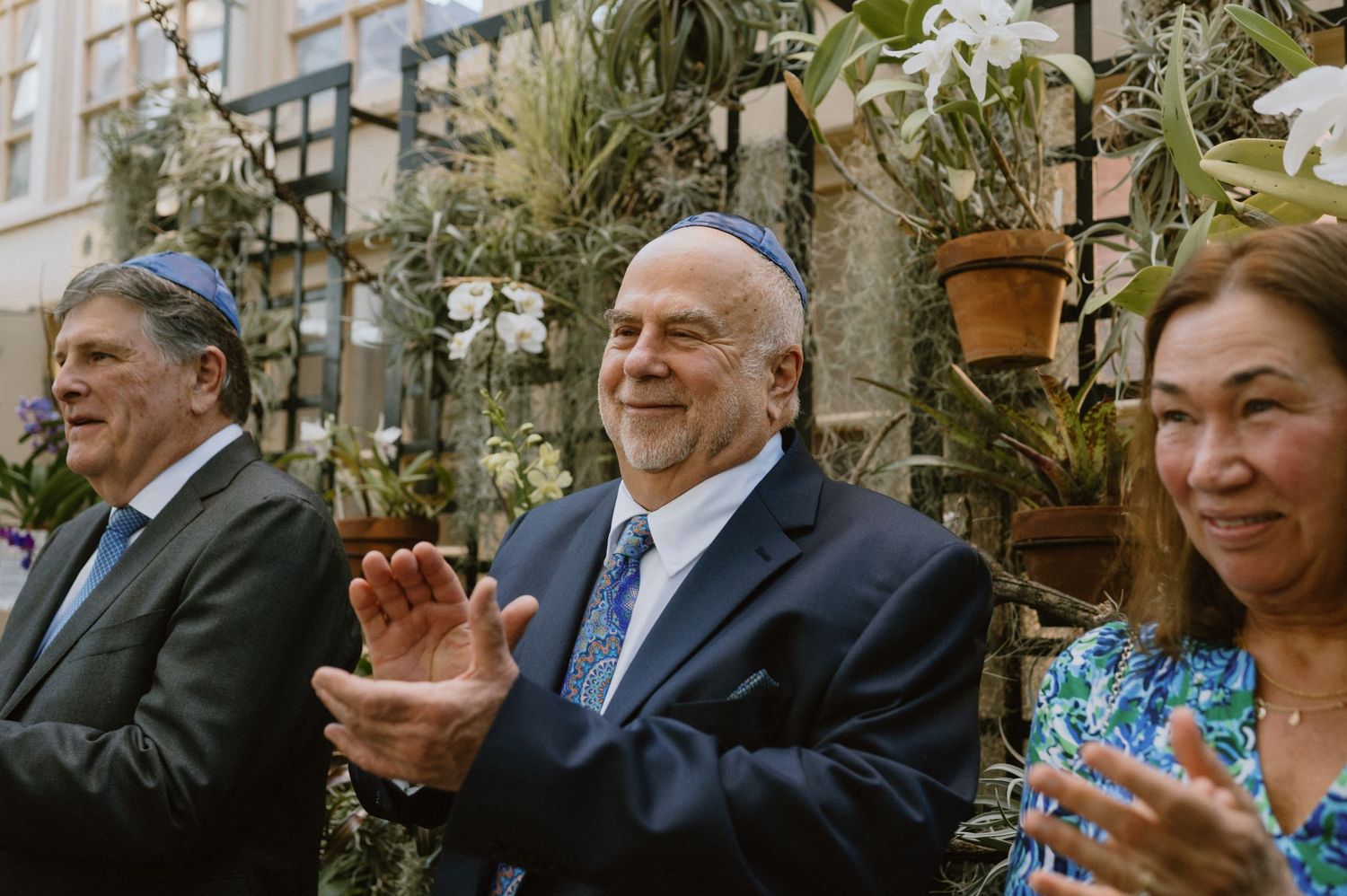 Wedding guests applaud during ceremony in tropical greenhouse setting.