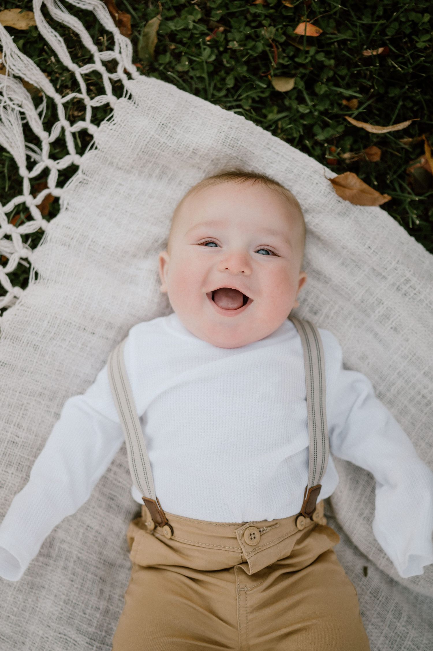 Happy baby wearing white top and tan suspenders lying on white blanket outdoors.