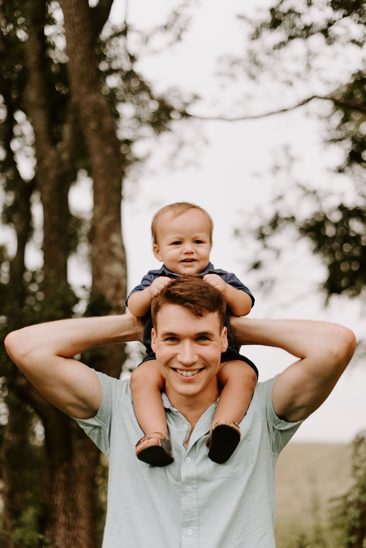 Baby sits on parent's shoulders against blurred forest backdrop during outdoor photoshoot.