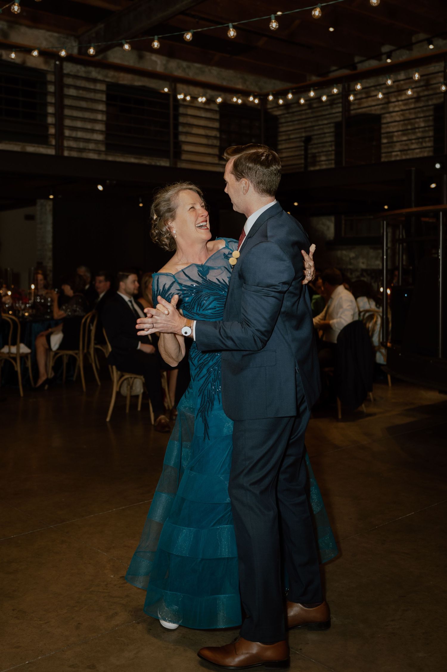 Couple dancing together in formal attire under string lights at an indoor venue.