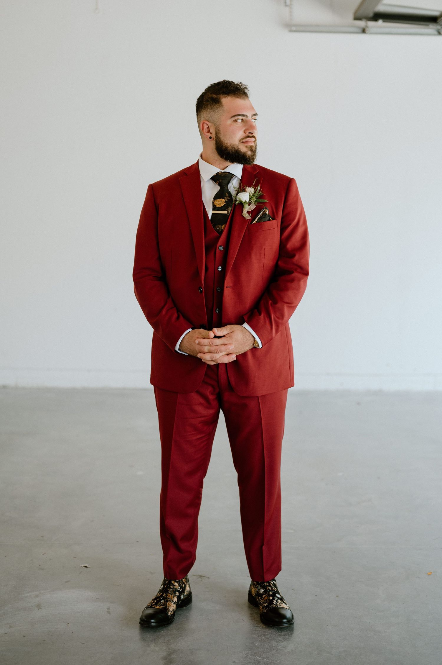 A groom in a burgundy three-piece suit stands confidently against a white wall.