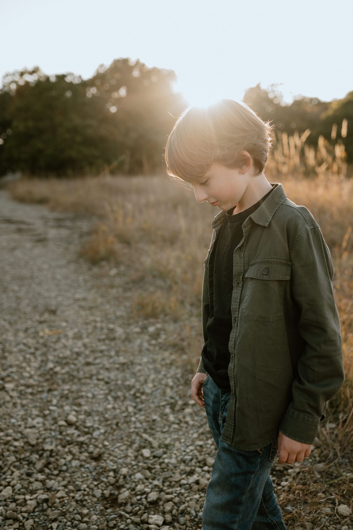 A backlit silhouette shows a child in casual wear standing on a gravel path during sunset.