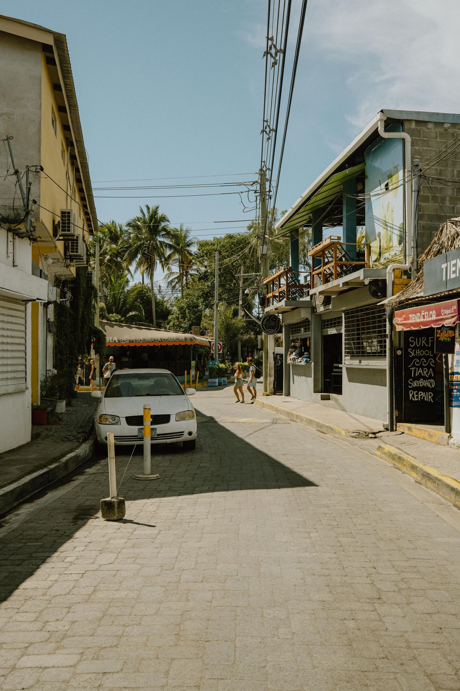 A quiet street in a tropical town with palm trees and parked cars between small buildings and shops.