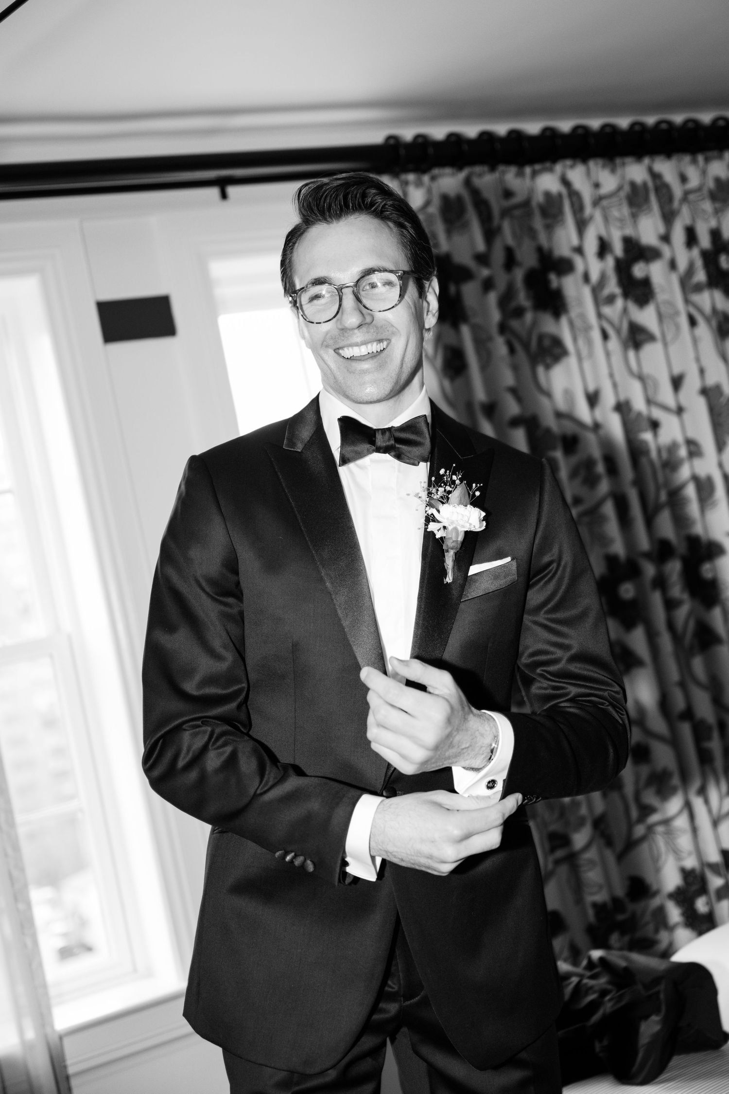 Black and white photo of person in glasses wearing a tuxedo with boutonniere and adjusting sleeves.