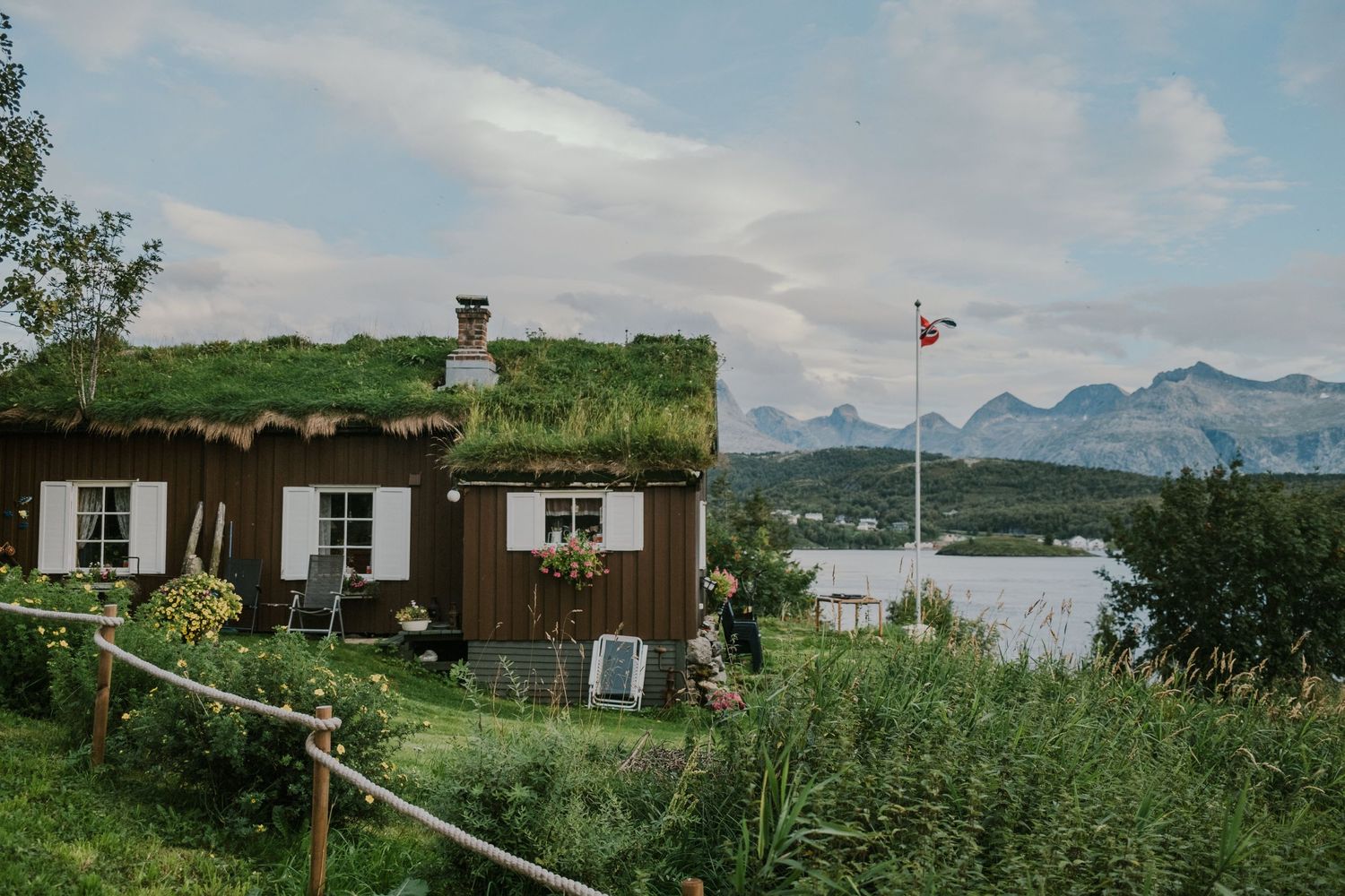 Rustic cabin with grass roof and Norwegian flag overlooking mountain fjord landscape.