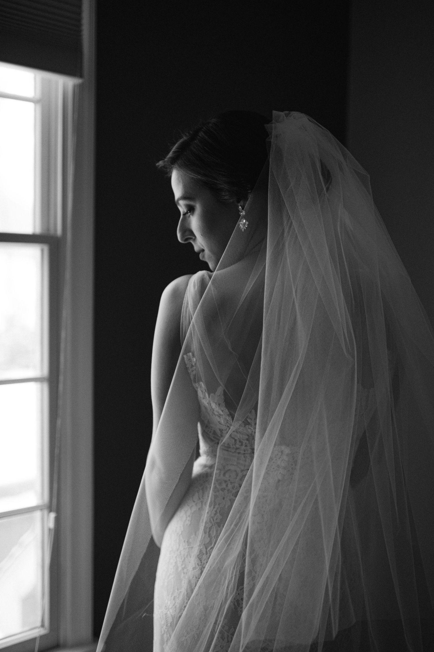 A silhouetted bride gazes out a window in a black and white portrait showing her veil and lace gown.