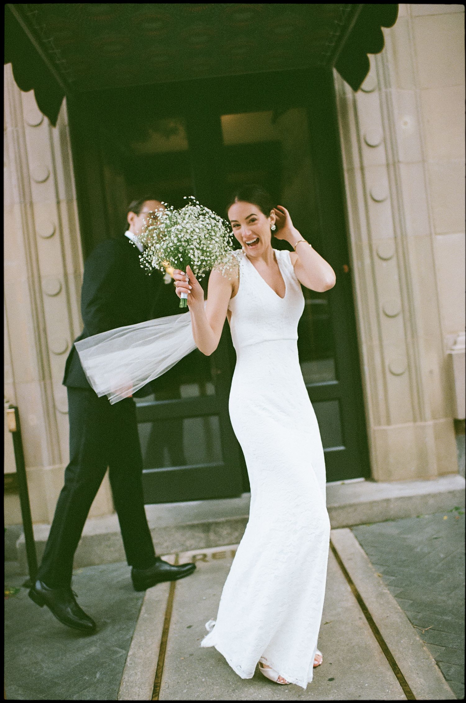 Joyful bride in sleek white dress laughs while holding bouquet of baby's breath.