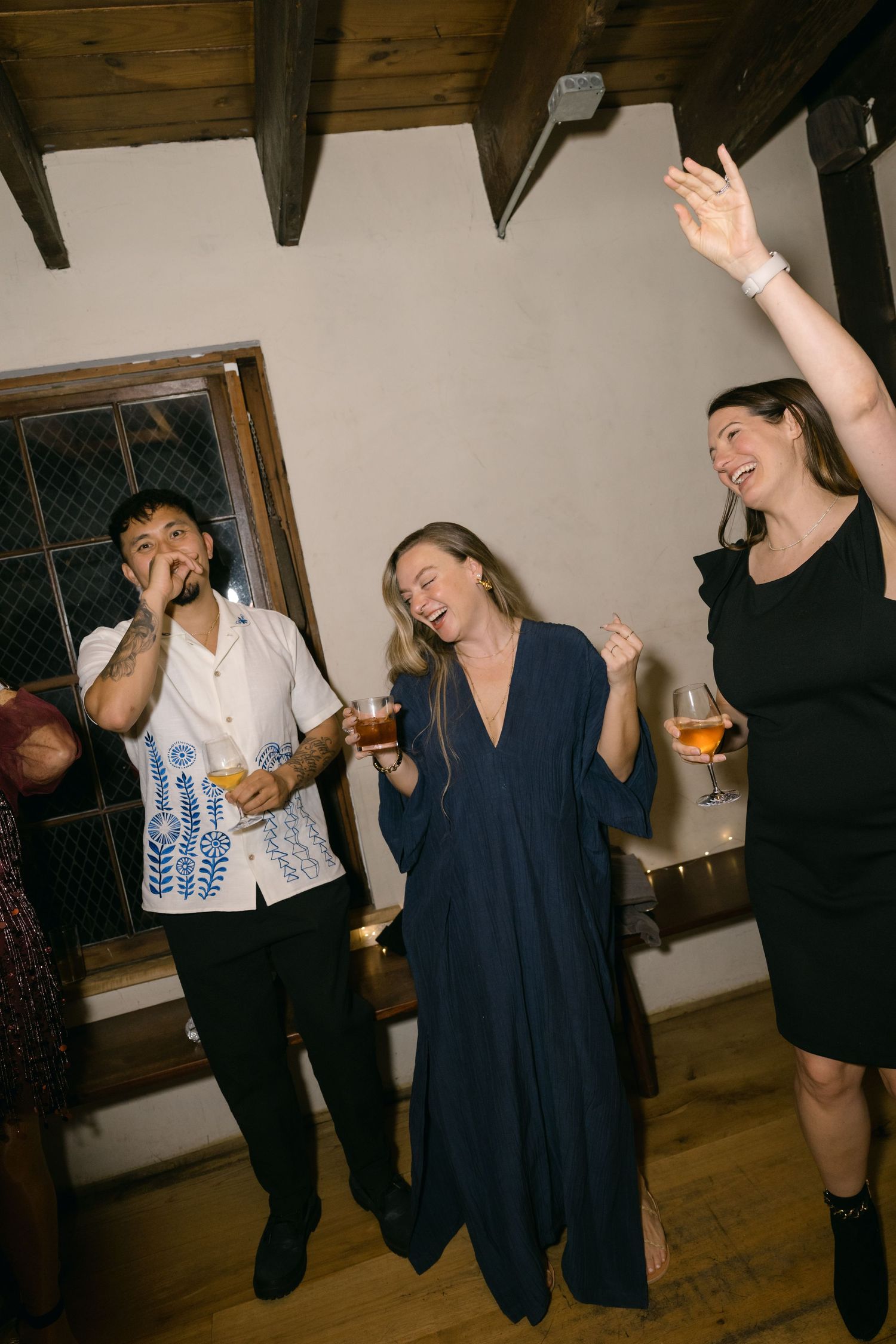 Three people dancing and singing together at a lively indoor party with wooden beams on the ceiling.