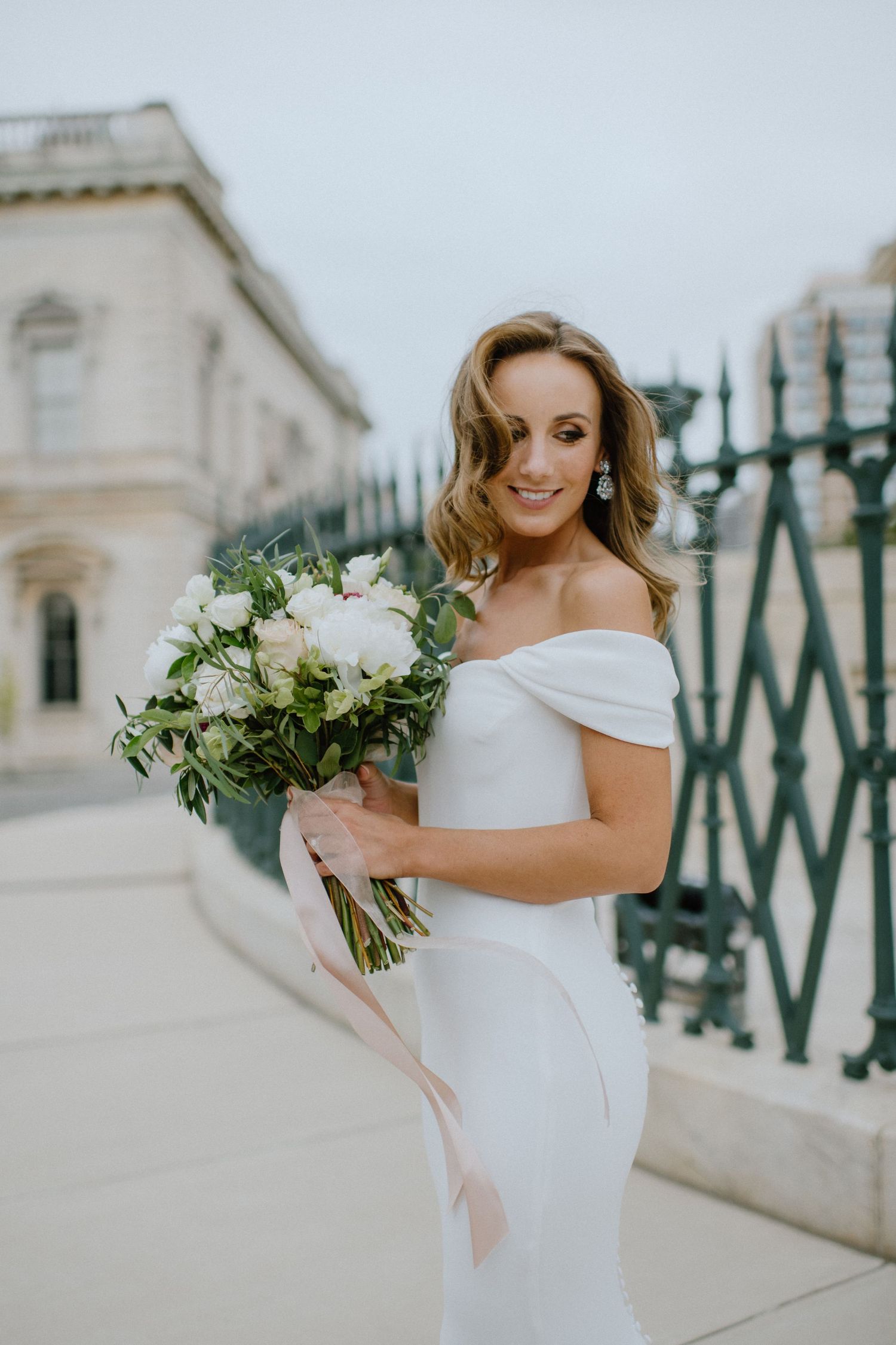 A bridal portrait showcasing an elegant white off-shoulder gown and natural bouquet against historic architecture.