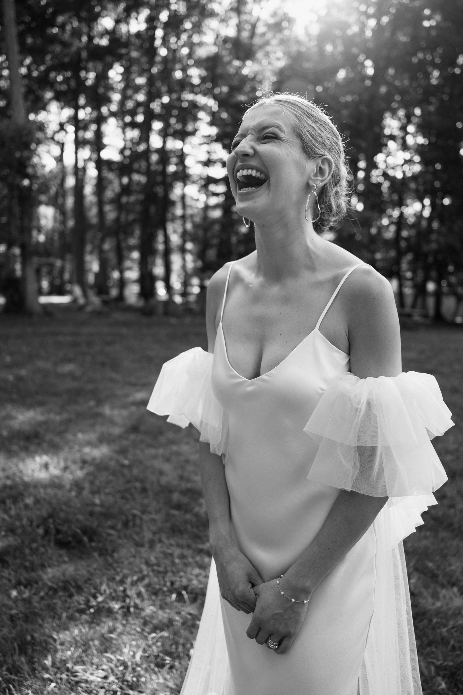 A joyful bride laughs candidly in her elegant off-shoulder wedding dress against a forest backdrop.