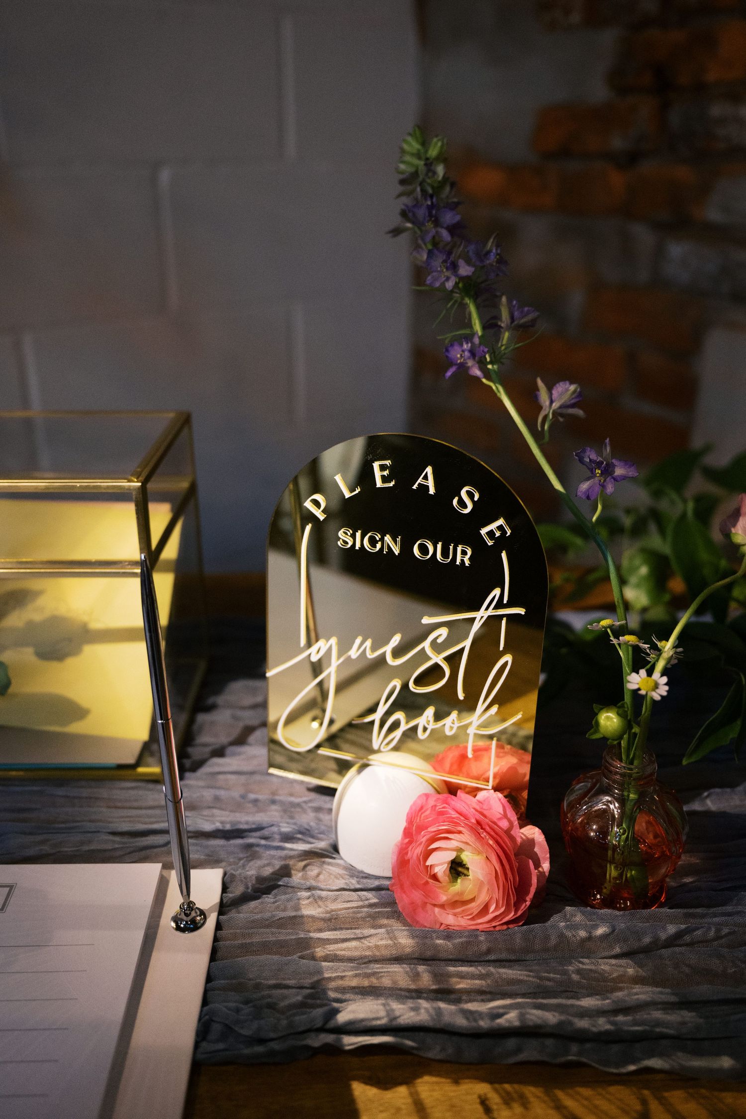An illuminated acrylic guest book sign with pink flowers and a glass lantern on a textured surface.