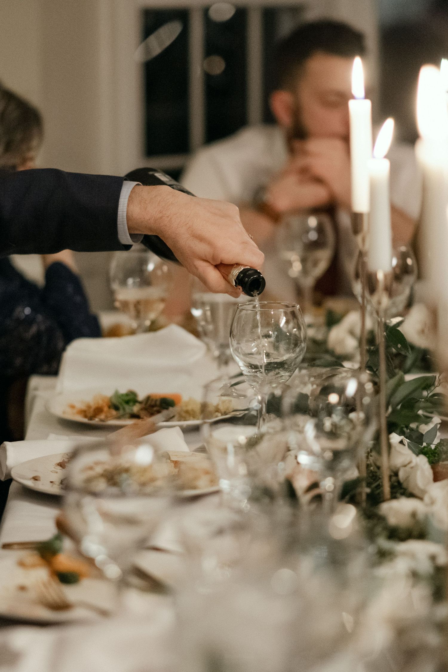 Wine being poured into glass at elegant candlelit wedding reception table with floral centerpieces.