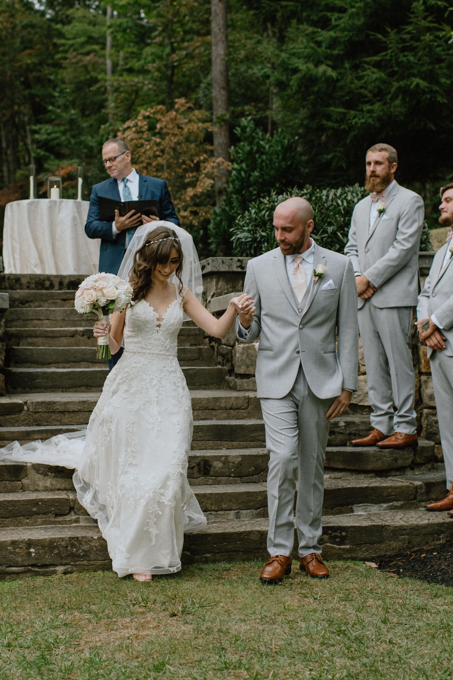 A couple descends stone steps at their outdoor garden wedding while attendants look on.