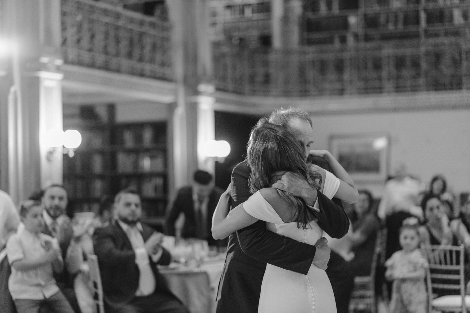 Black and white photo captures emotional embrace during wedding dance in ornate library setting.