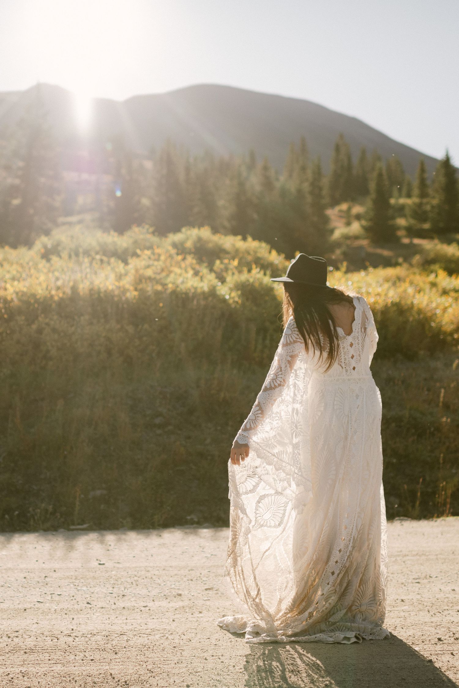 A person in a flowing lace dress stands on a mountain road at golden hour.