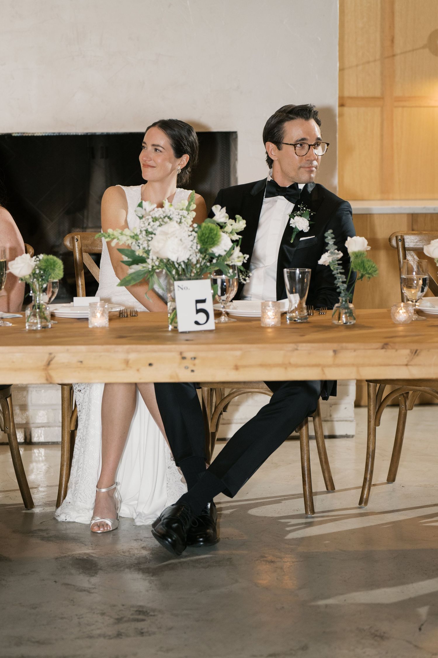 Newlyweds seated at rustic wooden table with white floral centerpieces and elegant table settings at reception.