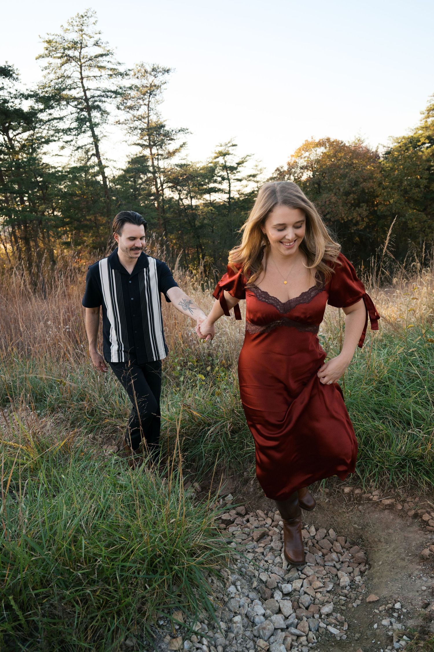 A couple walking together on a rocky trail during golden hour with pine trees and tall grass surrounding them.