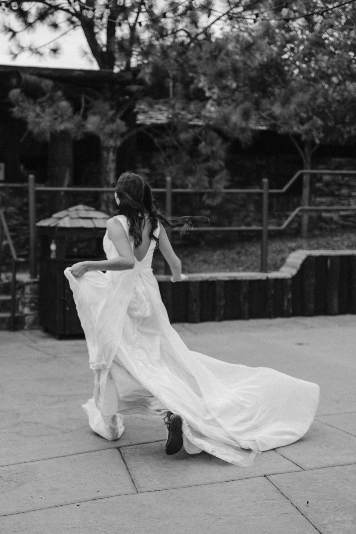 A black and white photo shows a flowing white wedding dress trailing behind someone wearing boots on a paved surface.