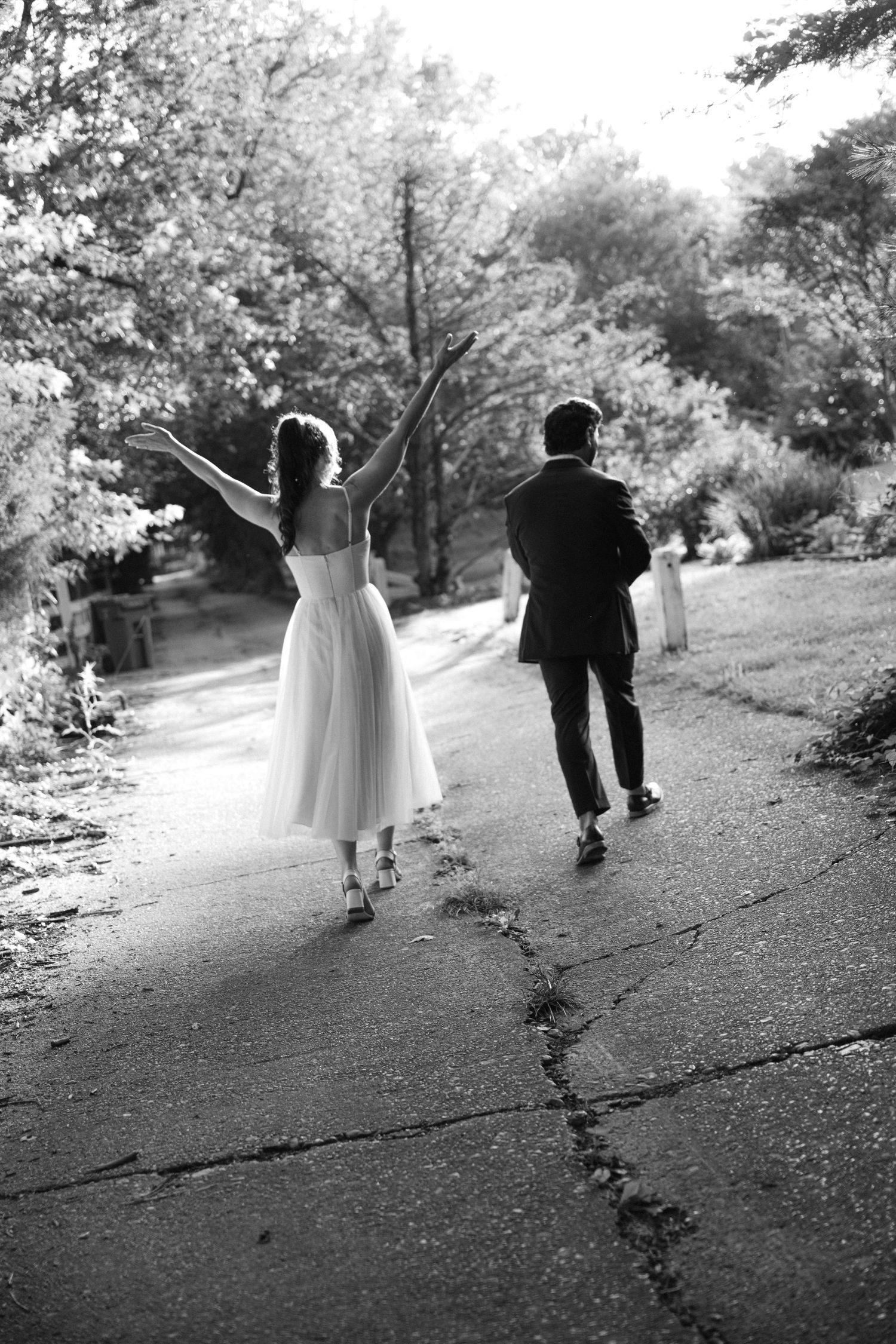 Black and white photo of a couple walking on a path, with person in dress joyfully raising arms while other walks ahead.
