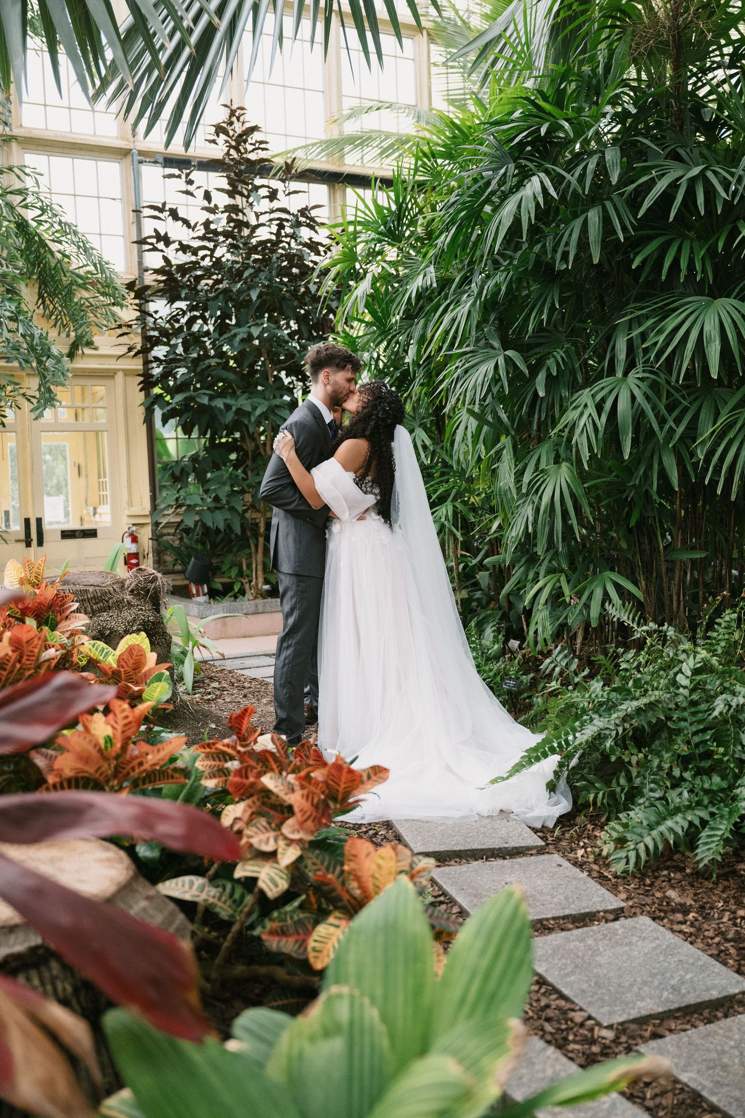 Bride and groom sharing a romantic kiss surrounded by lush tropical plants and colorful foliage in a greenhouse.