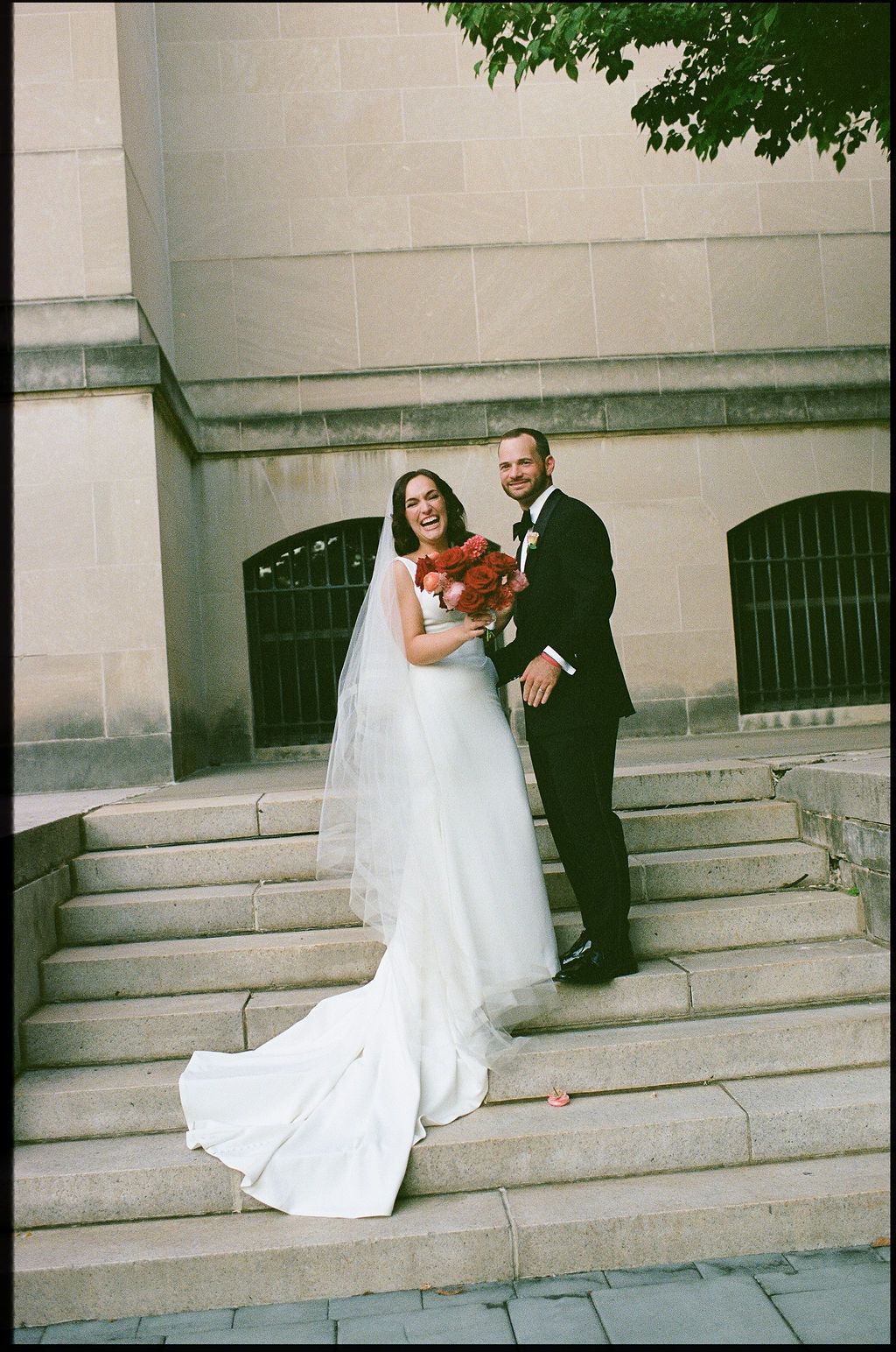 A bride in a flowing white gown and groom in a black suit pose together on stone steps outside a building.