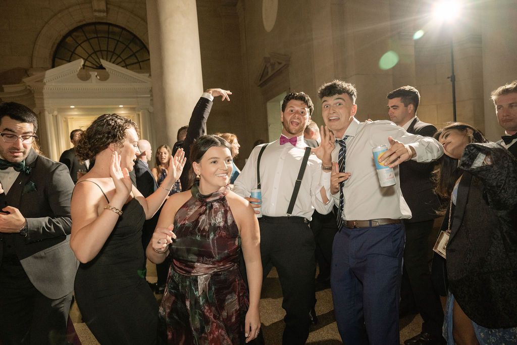 Wedding party guests dancing and celebrating on the dance floor at an indoor reception venue with bright lighting.
