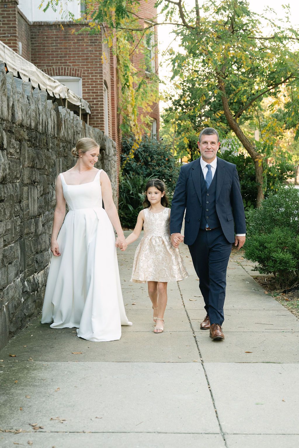 A wedding party walks along a tree-lined sidewalk next to a stone wall on a sunny day.