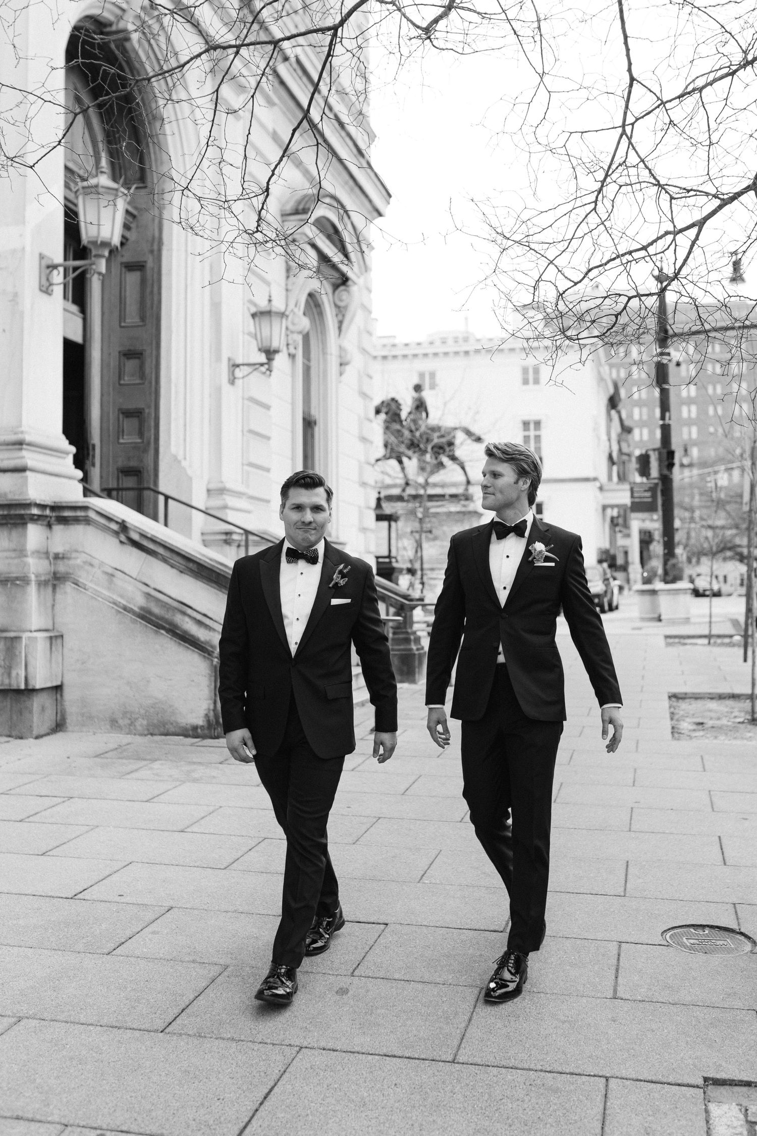 Men in formal black tuxedos walking along a stone pathway outside a grand building.