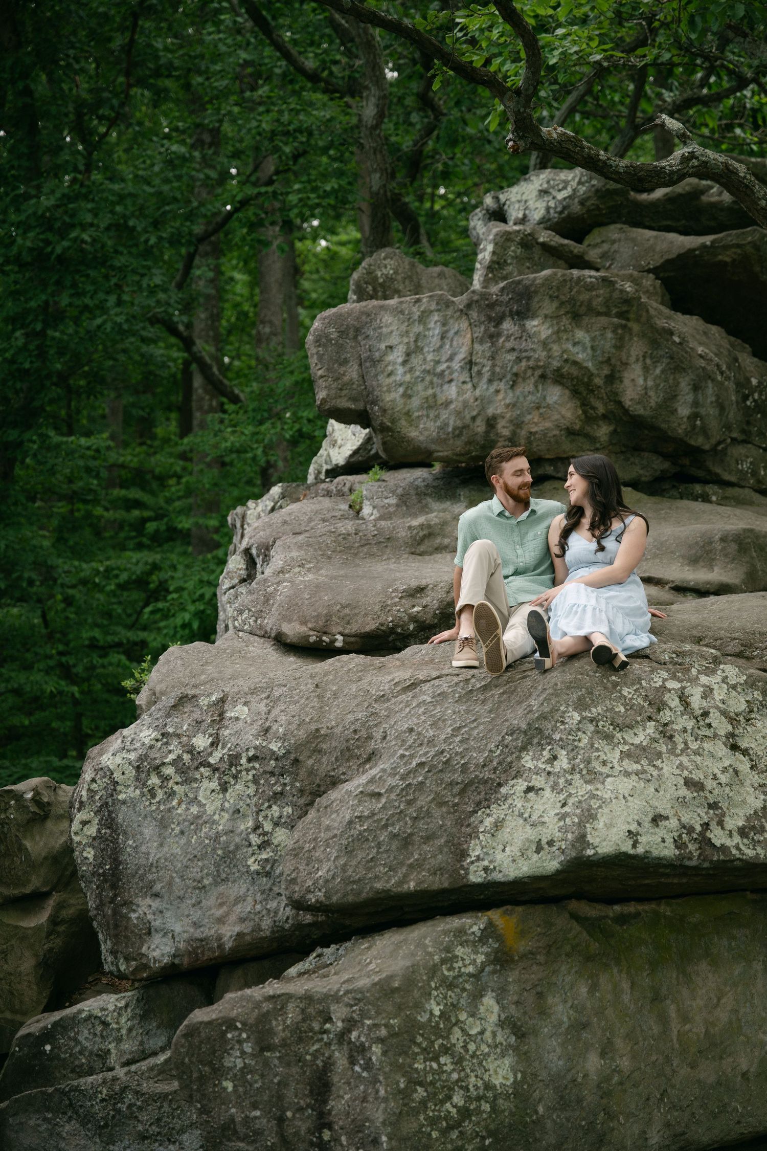 A couple relaxes on a large rocky outcrop surrounded by green forest.