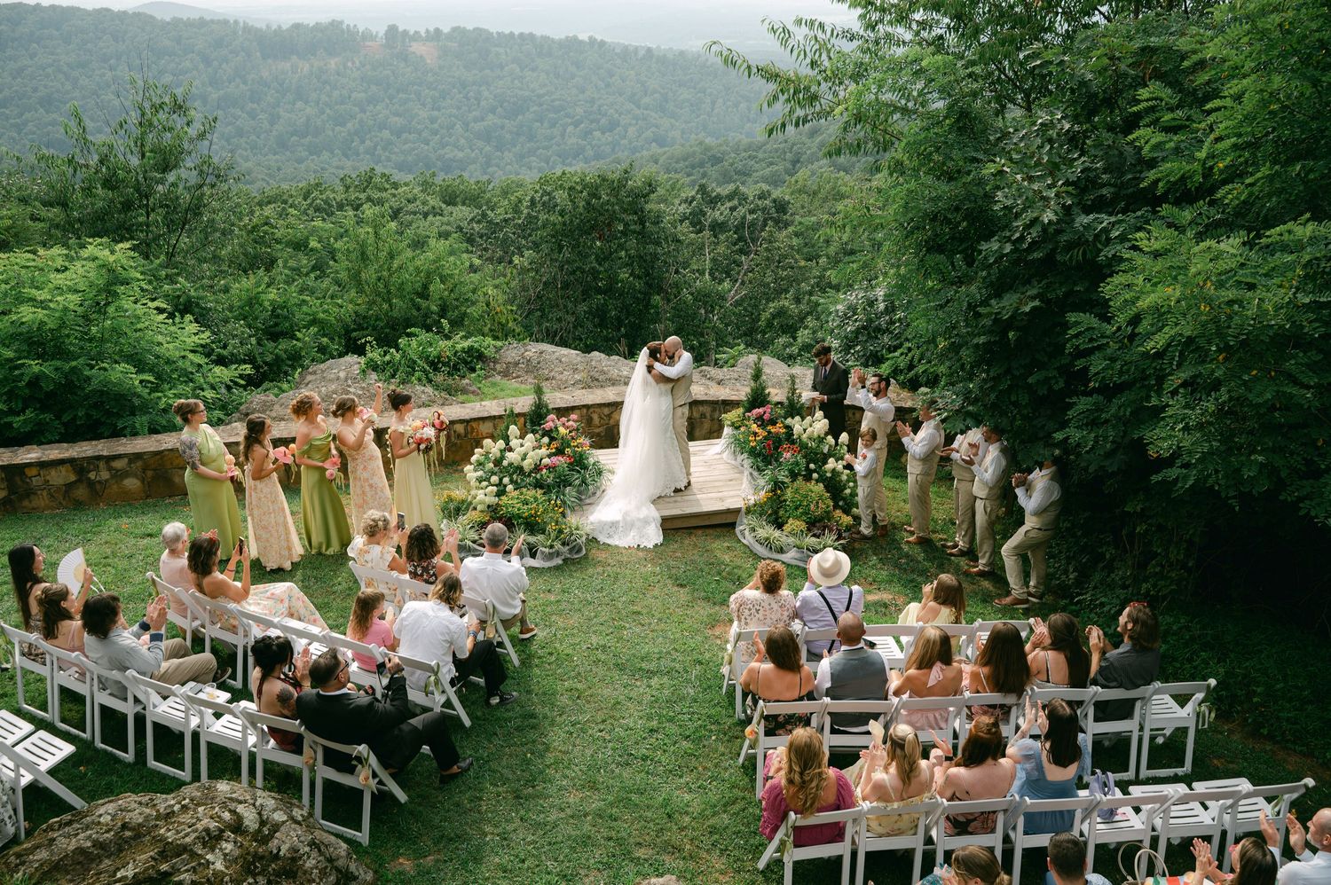 Outdoor wedding ceremony on a stone terrace overlooking lush green forest, with guests seated on white chairs facing the altar.
