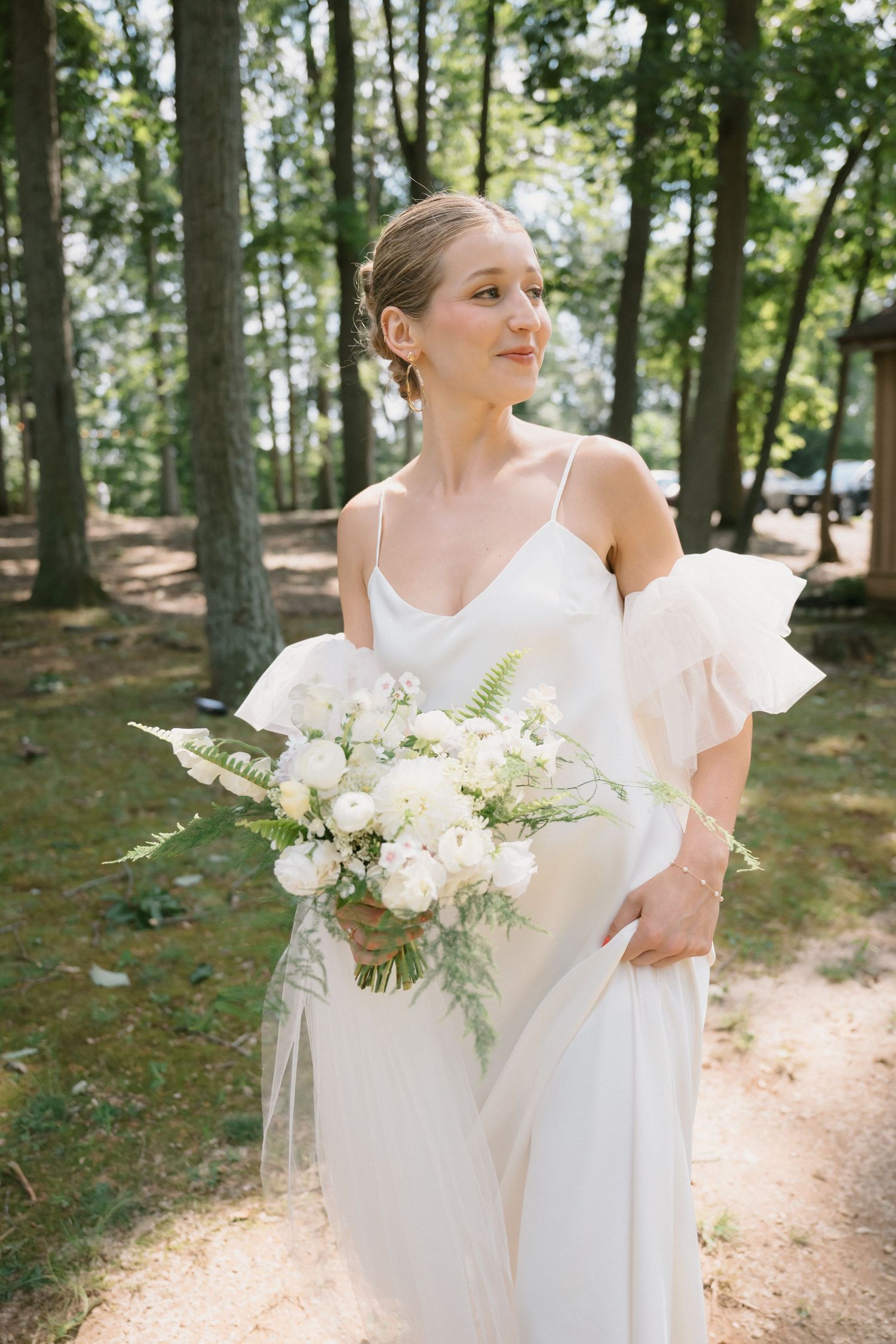 A bride holds a white floral bouquet while standing on a wooded path in an elegant dress.