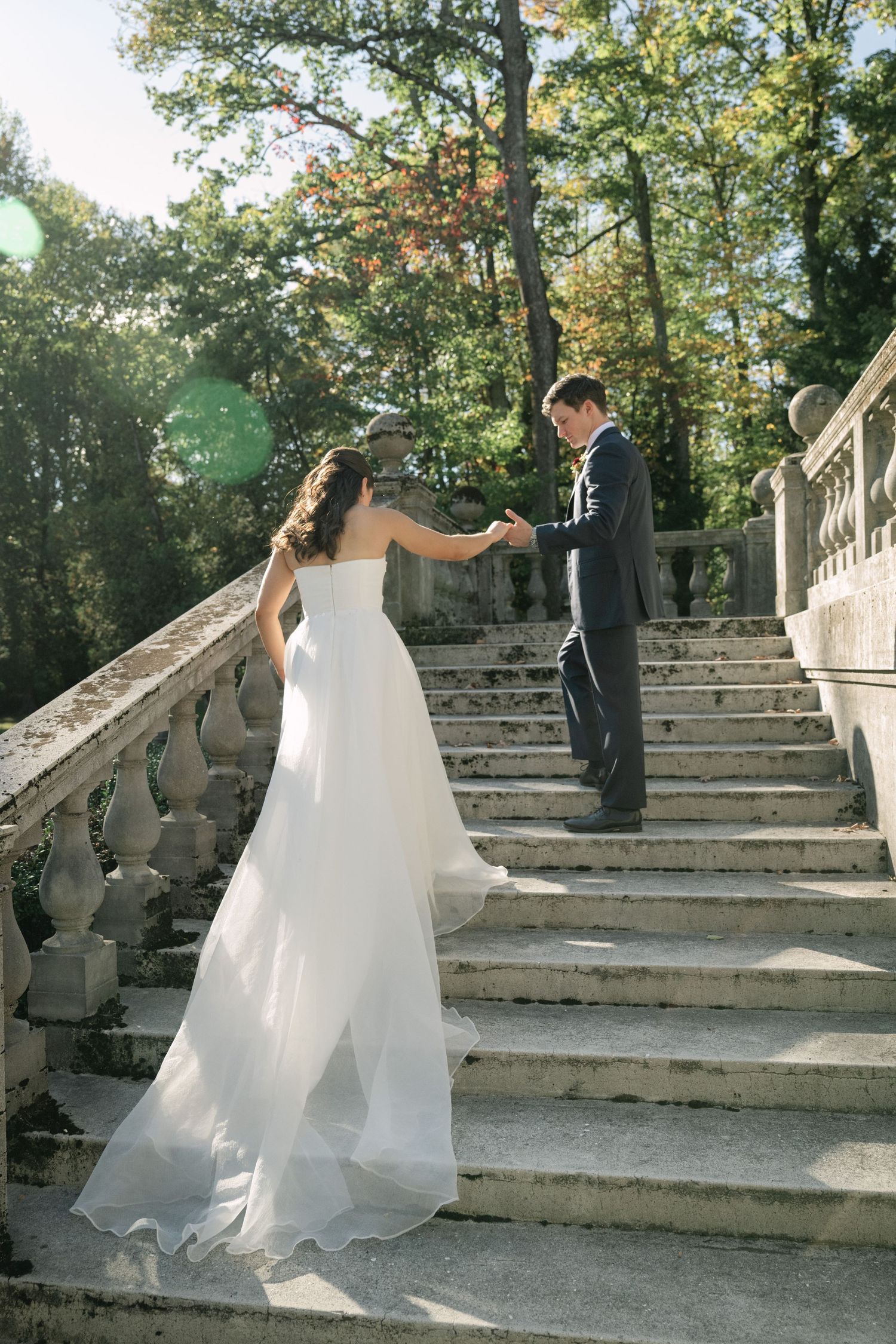 Bride in white gown holds groom's hand ascending elegant stone staircase surrounded by lush green trees.