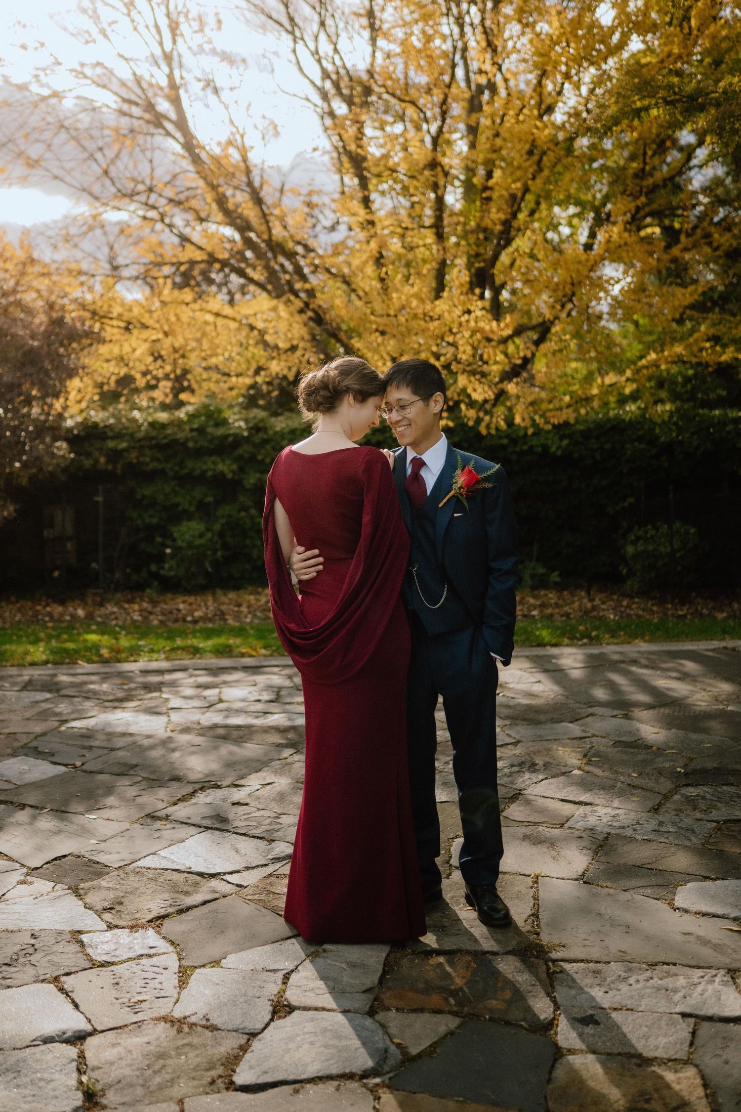 Couple in elegant fall wedding attire stand on stone patio surrounded by golden autumn trees.