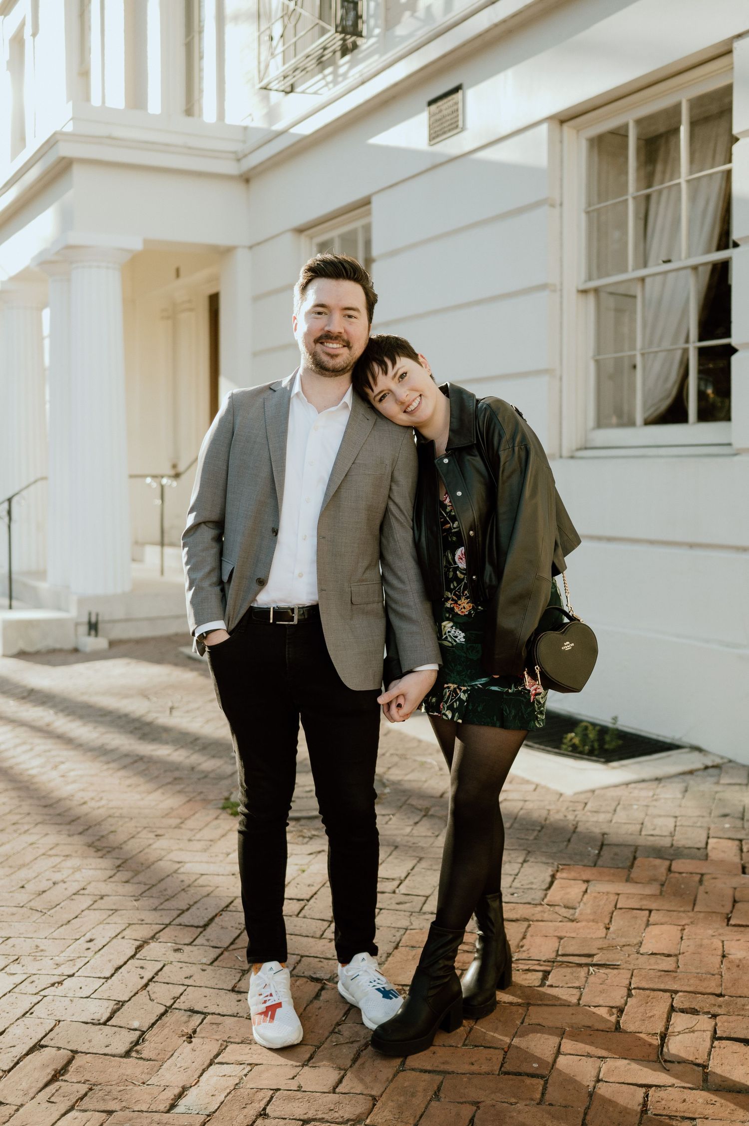 Couple posing together outside a white building, man in gray blazer and woman in floral dress leaning on his shoulder.