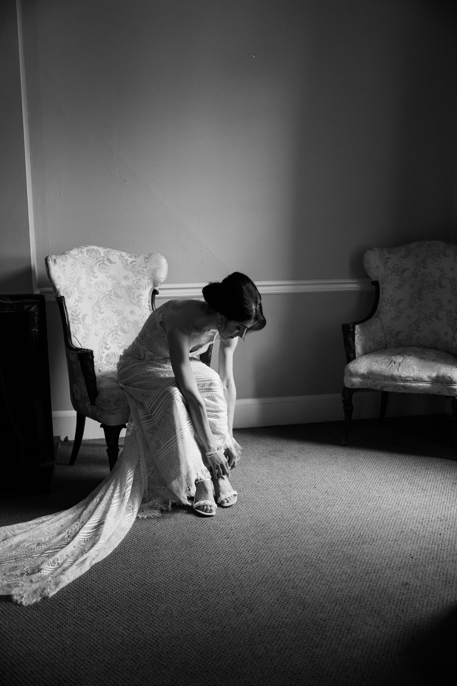 Black and white photo of a wedding dress while putting on shoes in an elegant room.