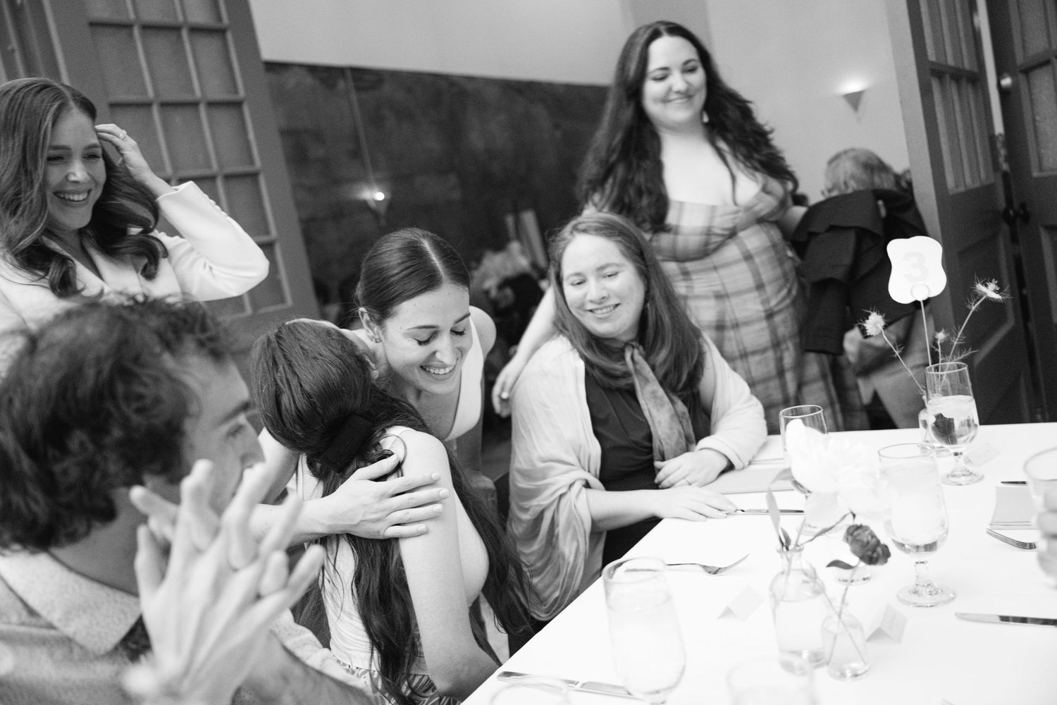 A candid black and white photograph of people laughing together around a dining table during a social gathering.