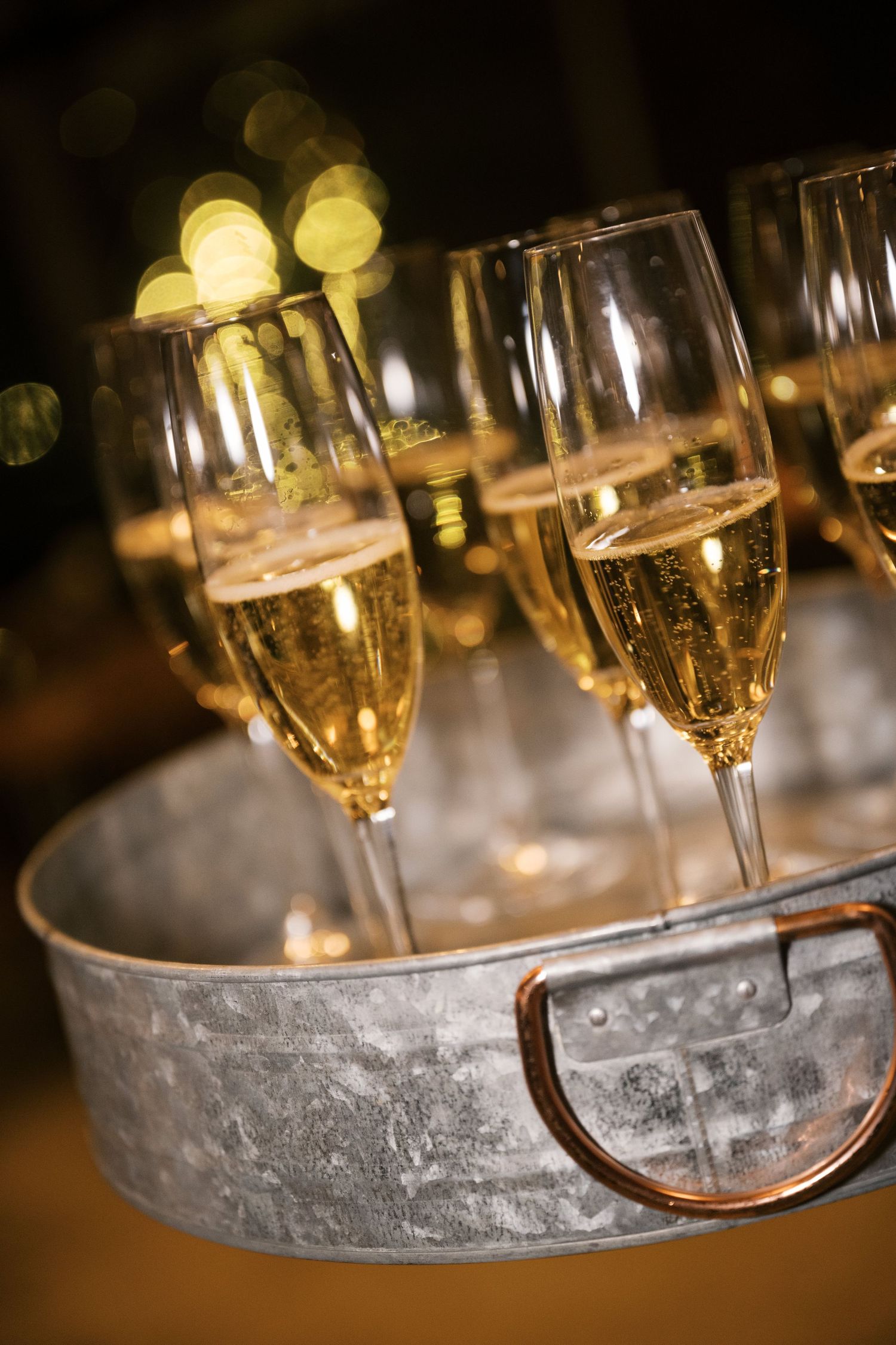 Champagne flutes filled with bubbly wine arranged in a silver serving tray with bokeh lights in background.