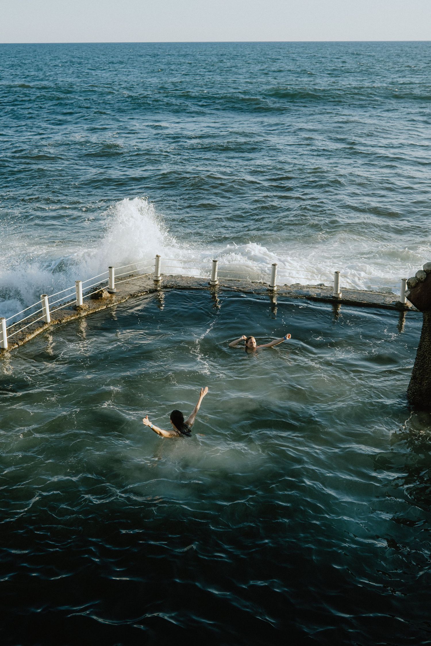 People swimming in an ocean pool protected by white posts as waves splash over the barriers.