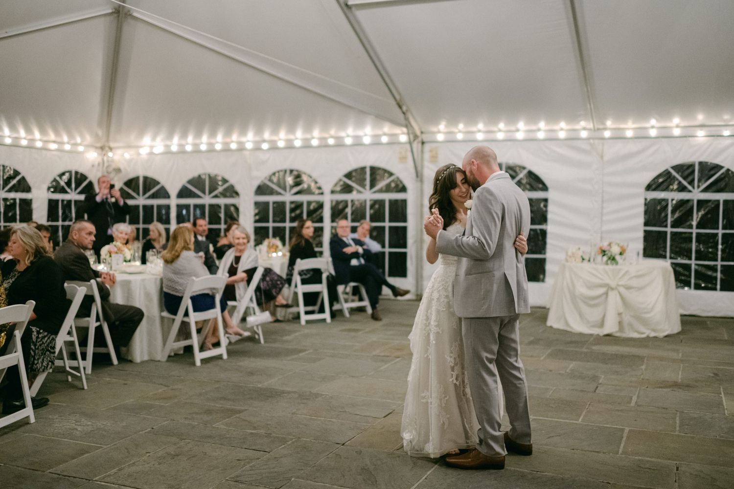 A couple shares their first dance under a white tent reception while guests watch from their seats at tables.