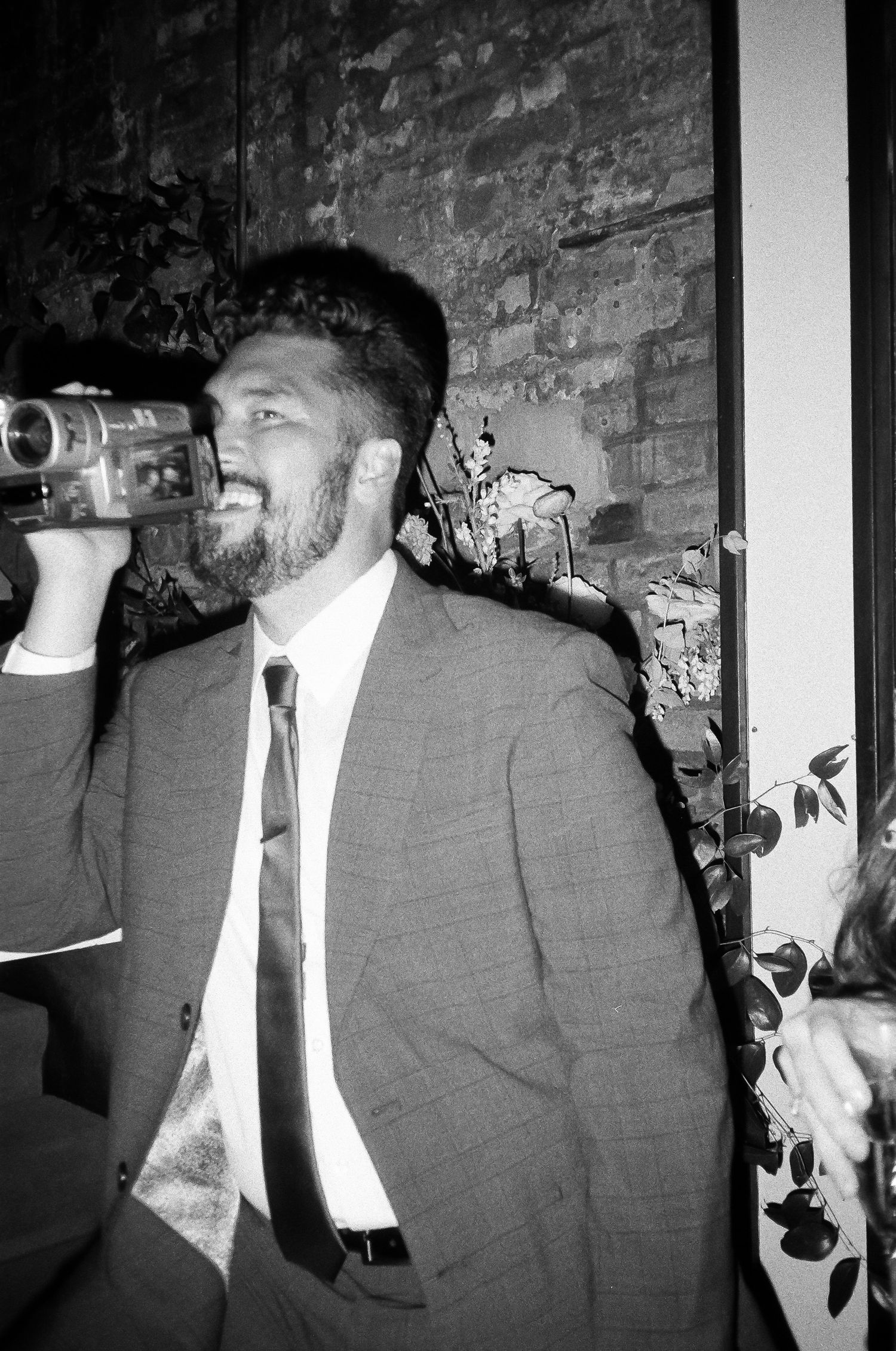 Man in suit jacket and tie using a video camera against brick wall background.