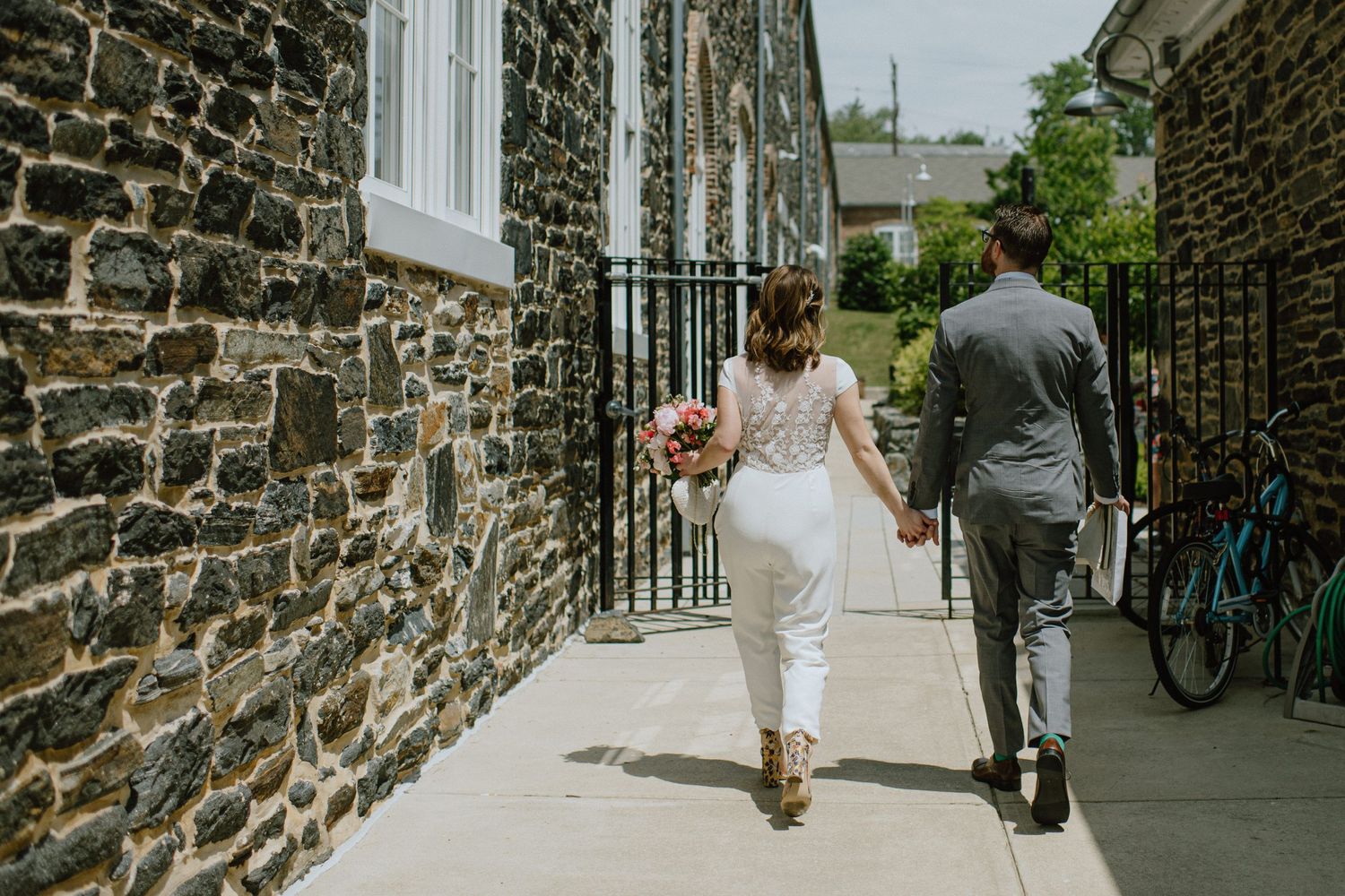 Couple in wedding attire walking hand in hand along a stone-walled pathway.