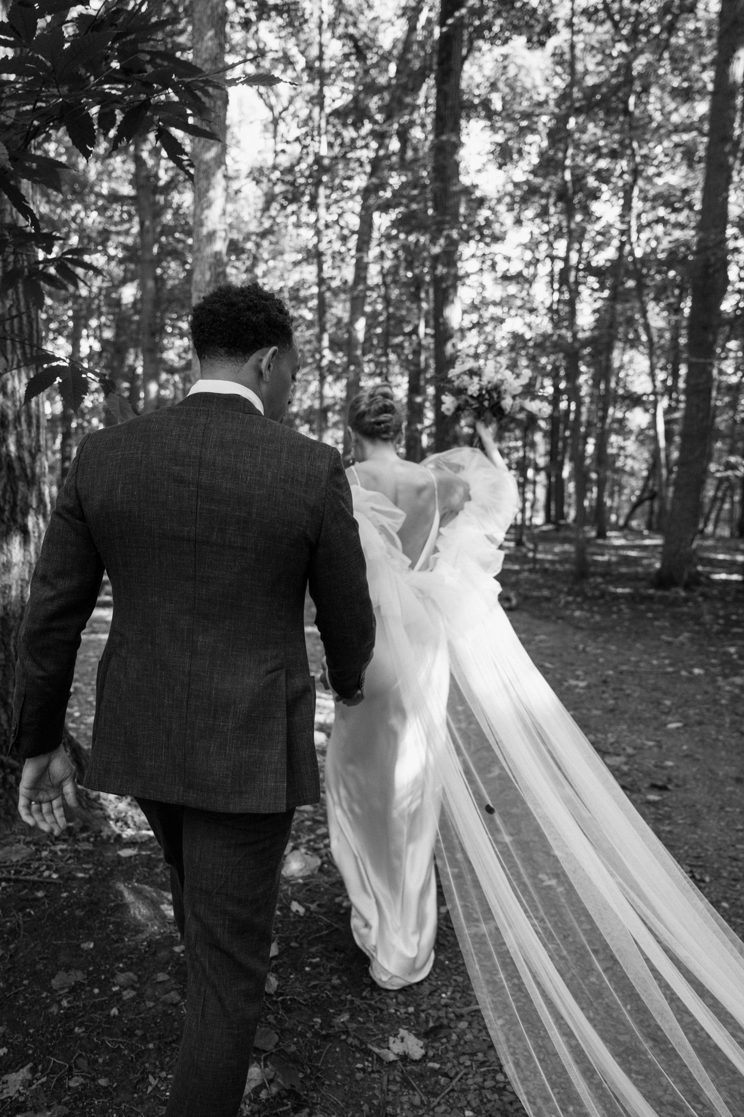 A flowing wedding veil trails behind as the couple walks through a wooded area.