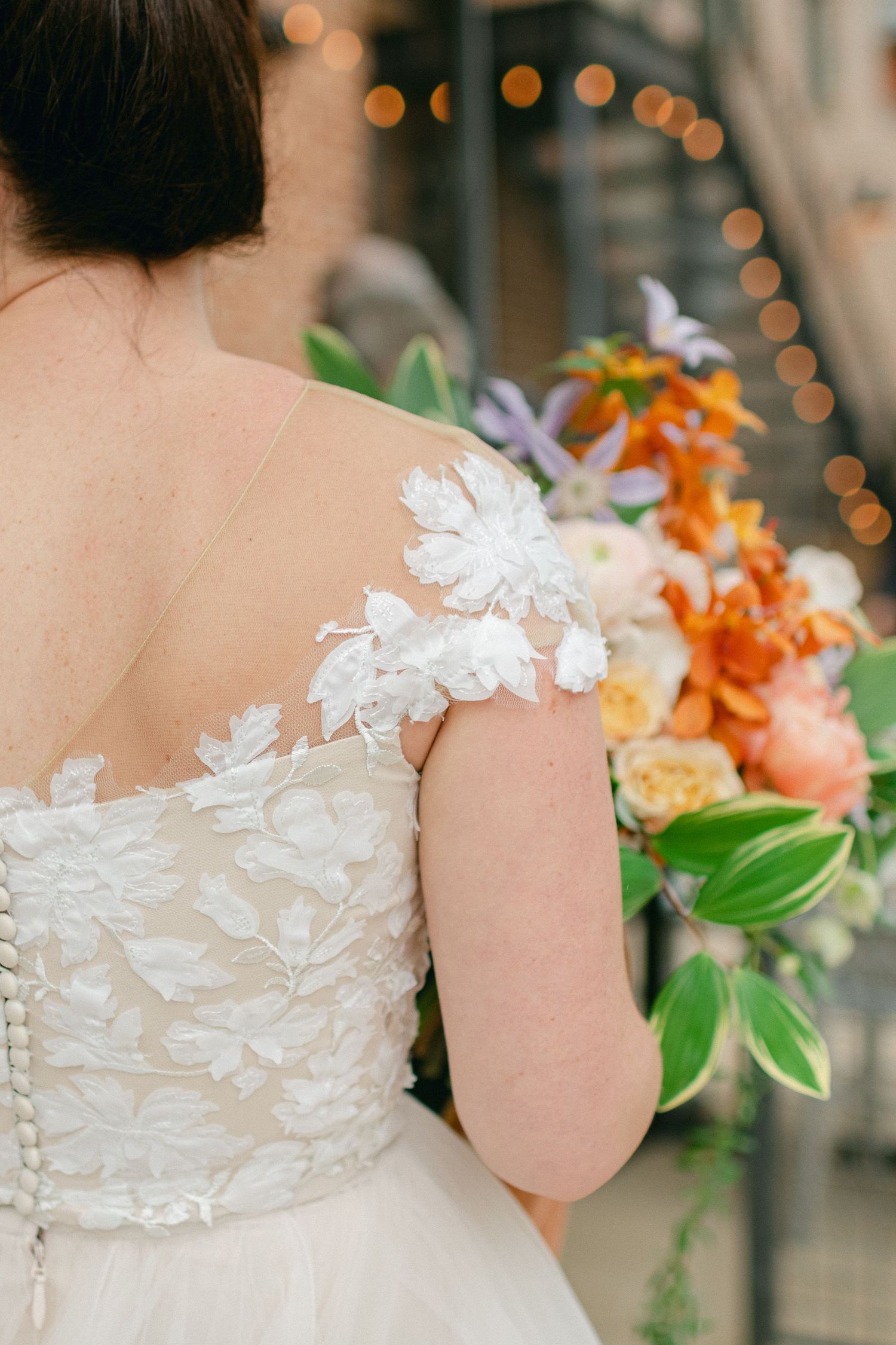 Close-up detail of a wedding dress with delicate floral appliques and a colorful bouquet.