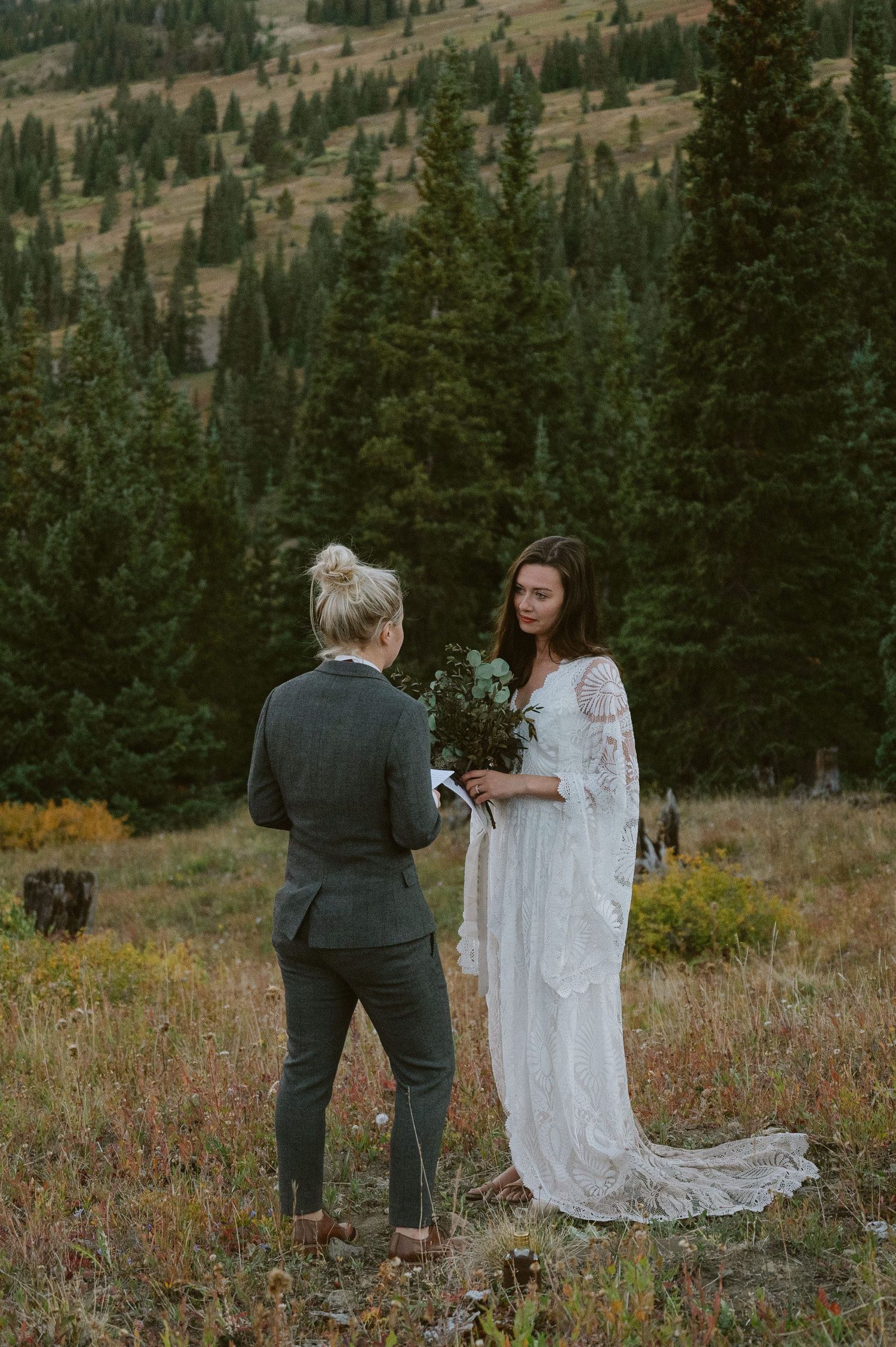 Mountain wedding ceremony with lace dress and suit against evergreen forest backdrop.