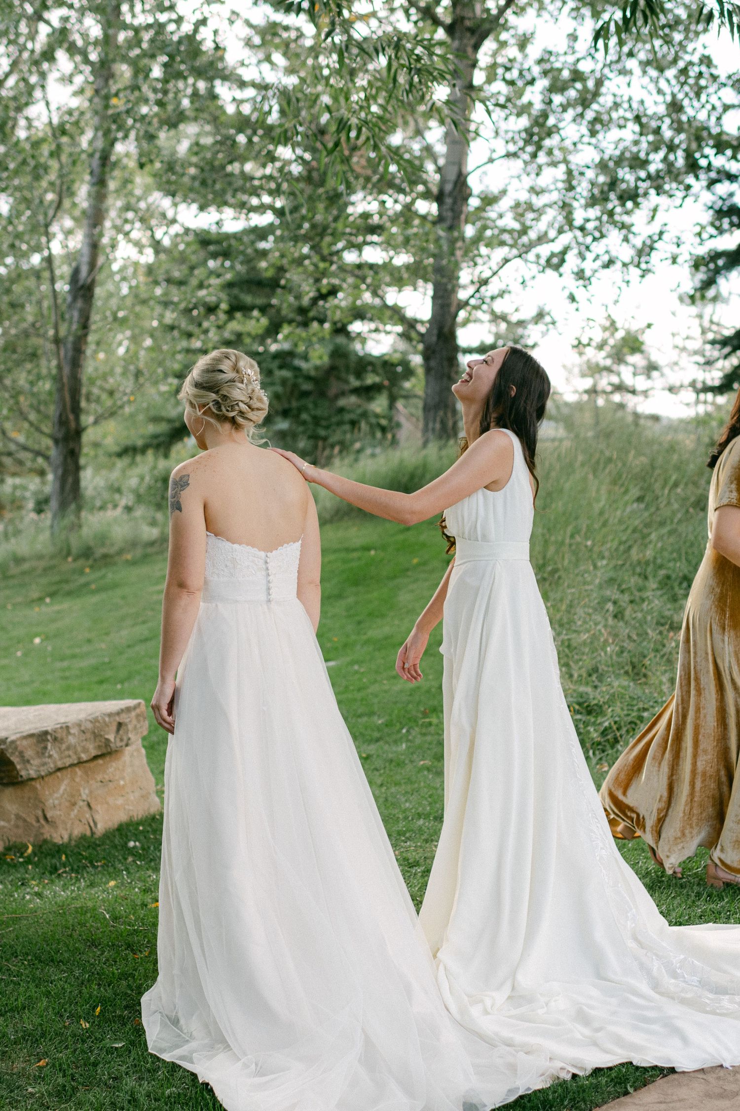 Two women in white dresses sharing a moment on grassy hillside.