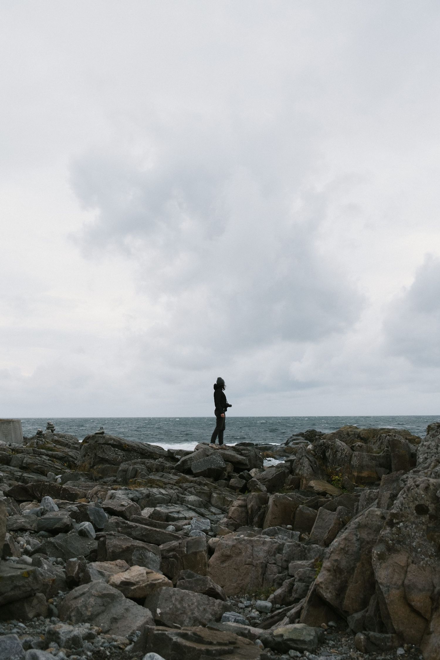 Silhouette of person standing on rocky coastline overlooking ocean under moody sky.