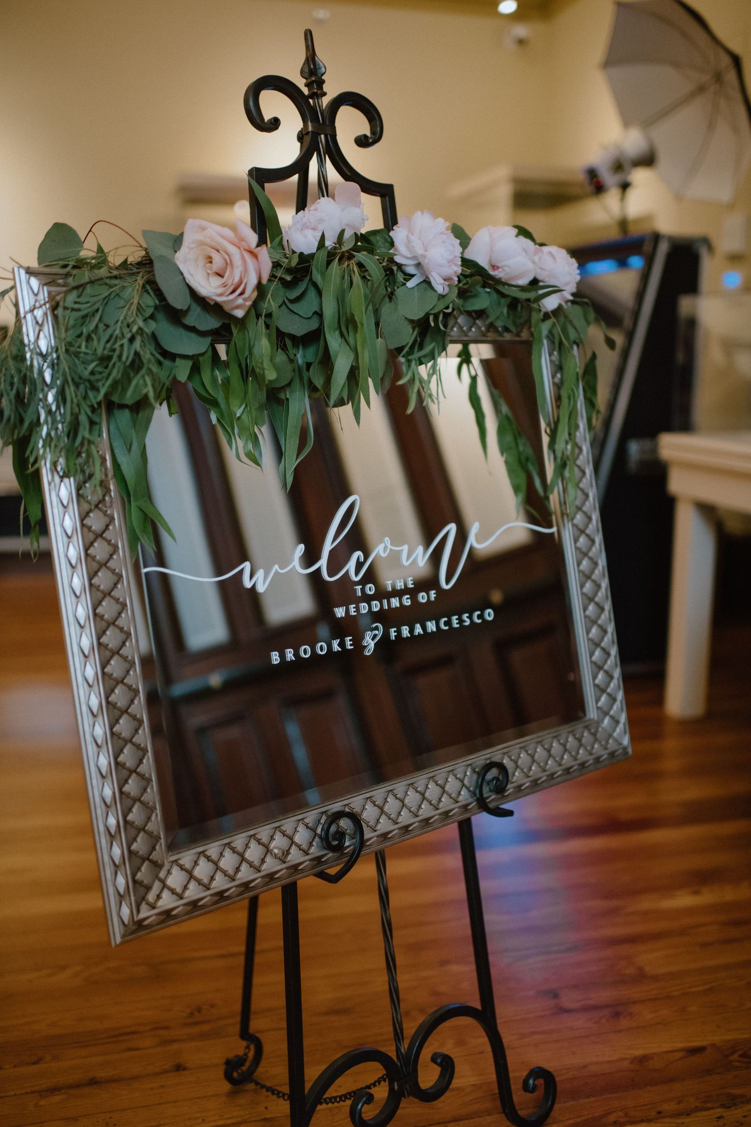 Ornate welcome sign with roses and greenery in a decorative frame on an easel.