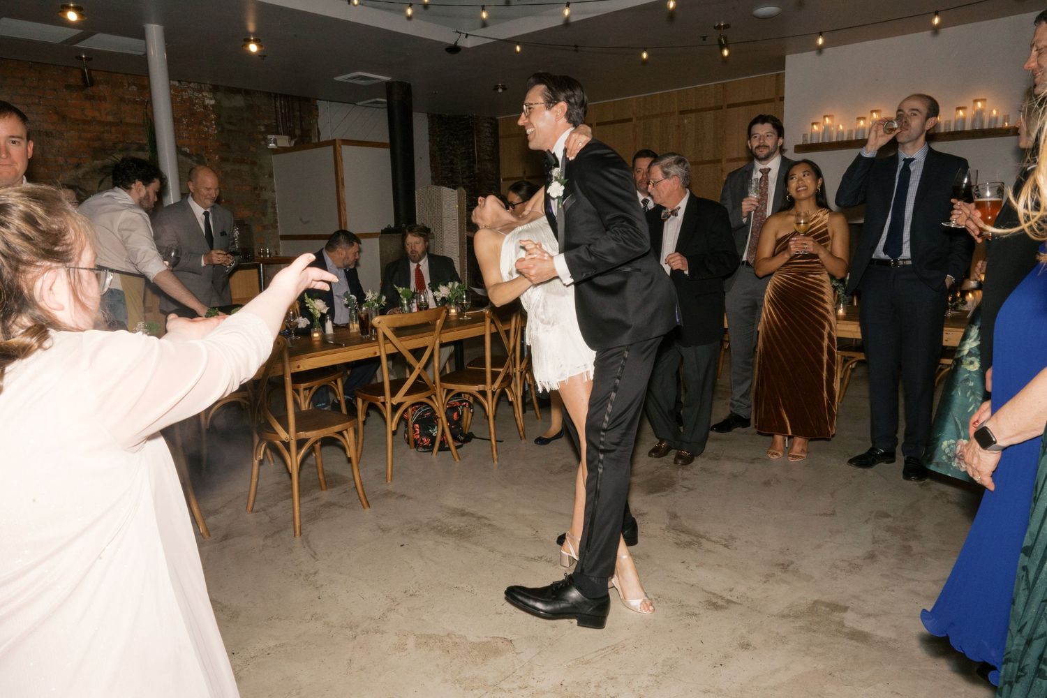 Guests watching a couple dance at a reception with rustic brick walls and wooden chairs in the background.