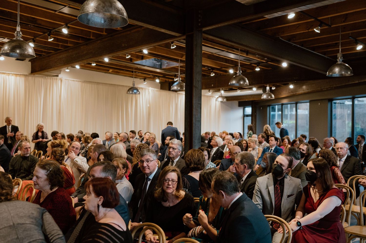 Large crowd seated in rows of wooden chairs at an indoor event venue.