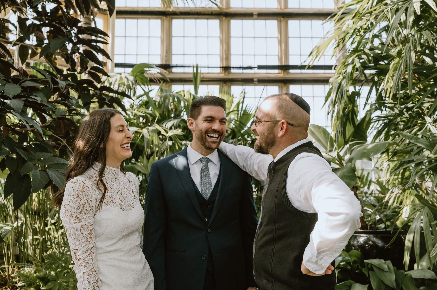 Candid moment of laughter between couple and officiant during greenhouse wedding ceremony.